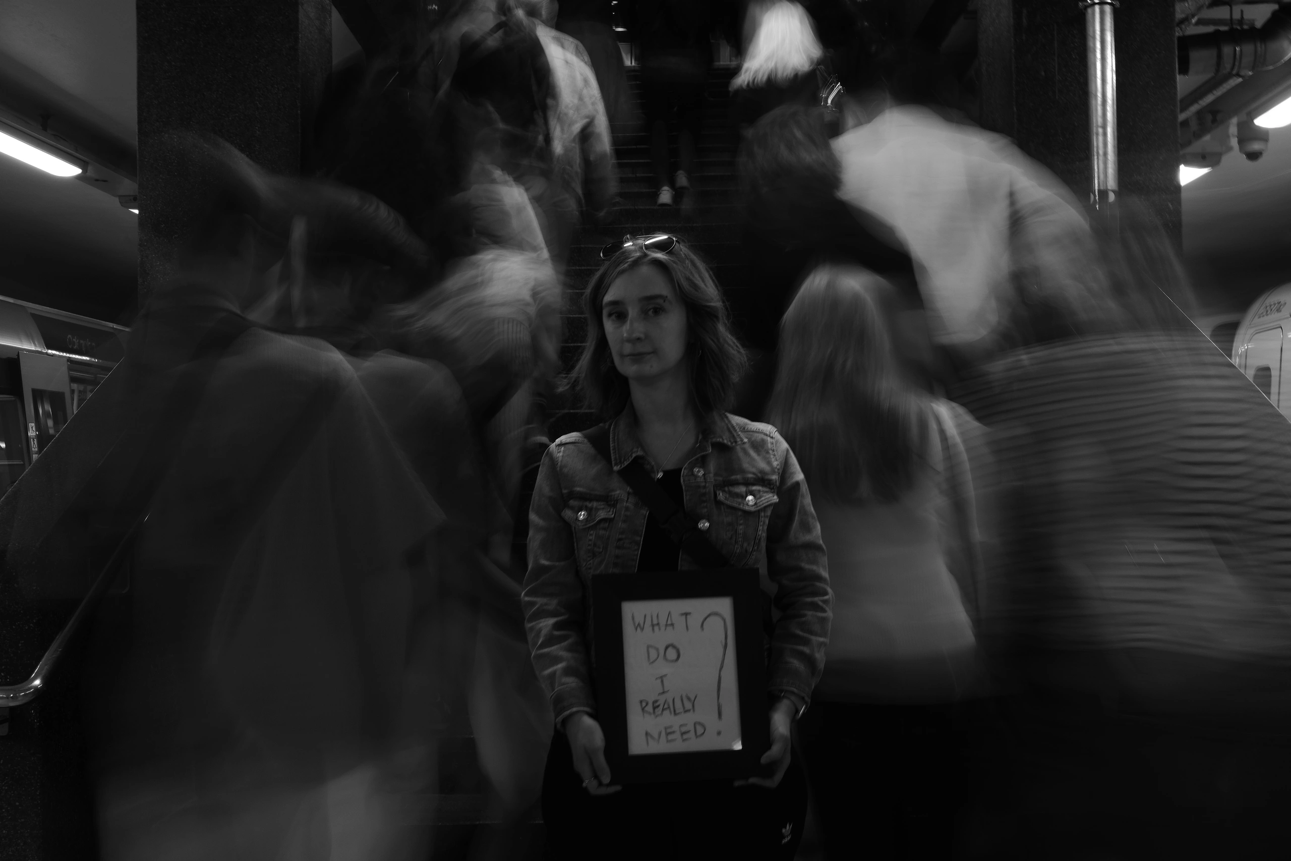Woman holding a sign reading What do I really need as blurred commuters pass
